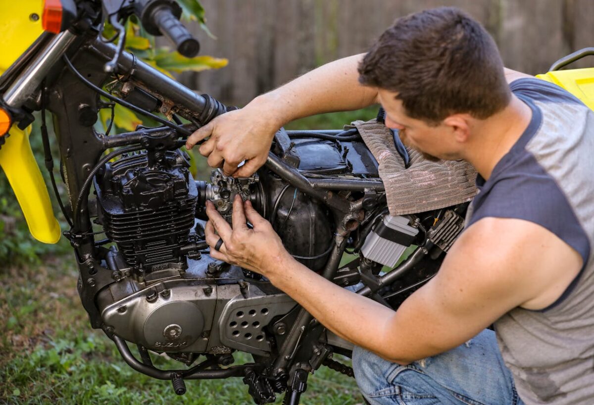 Mechanic in sleeveless shirt working on motorcycle engine outdoors.