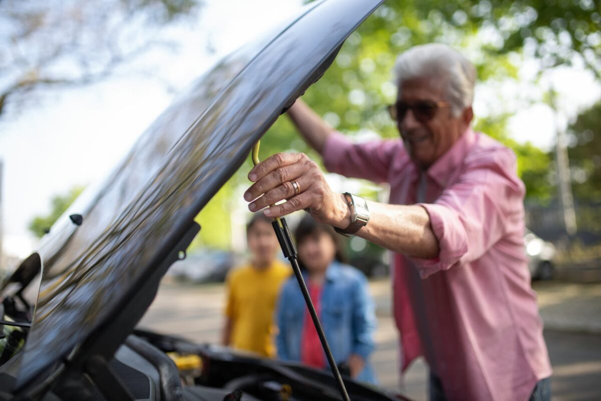 A senior man adjusts his car's hood strut while his grandchildren watch on a sunny day.
